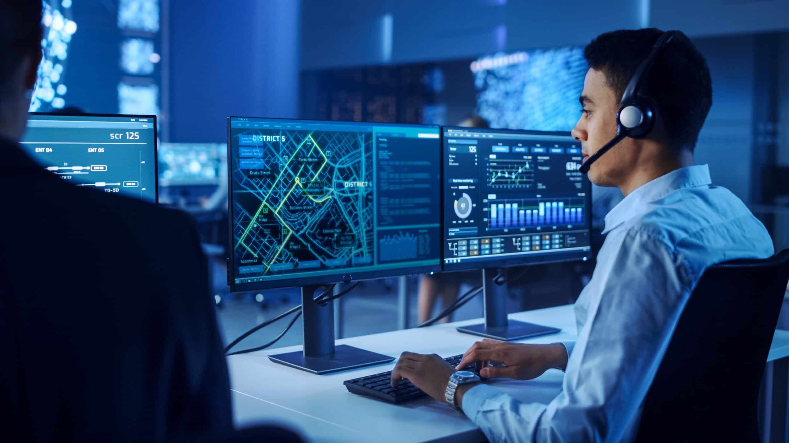 Confident Male Data Scientist Works on Personal Computer Wearing a Headset in Big Infrastructure Control and Monitoring Room. Map Surveillance of People Walking on City Streets